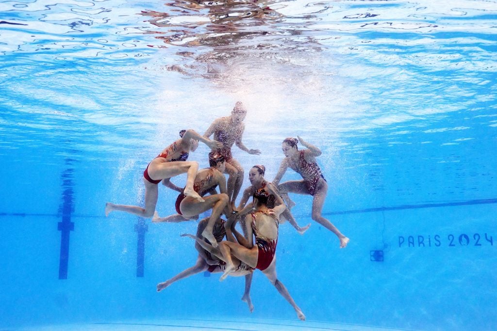 Adam Pretty used an underwater robotic camera to get this photo of China competing in the Team Acrobatic Routine in the 2024 Paris Olympic Games Paris. Eight female swimmers in dark red bathing suits with gold sequins get into position underwater in a swimming pool for an acrobatic lift.