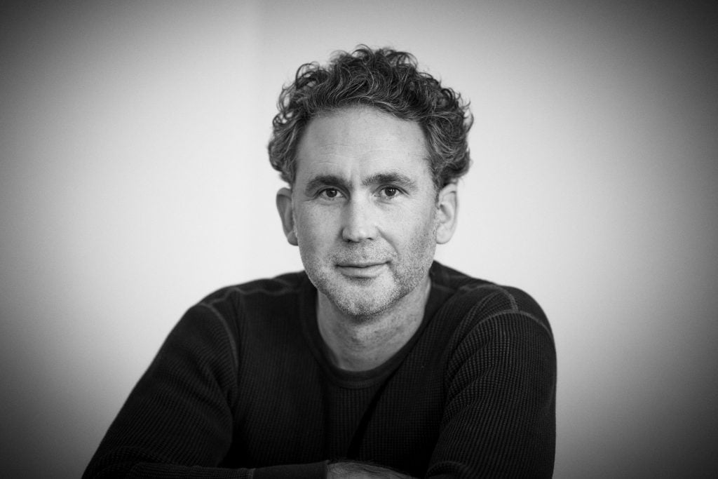 A black and white portrait of photographer Adam Pretty, a middle aged white man with dark curly hair wearing a dark sweater. 