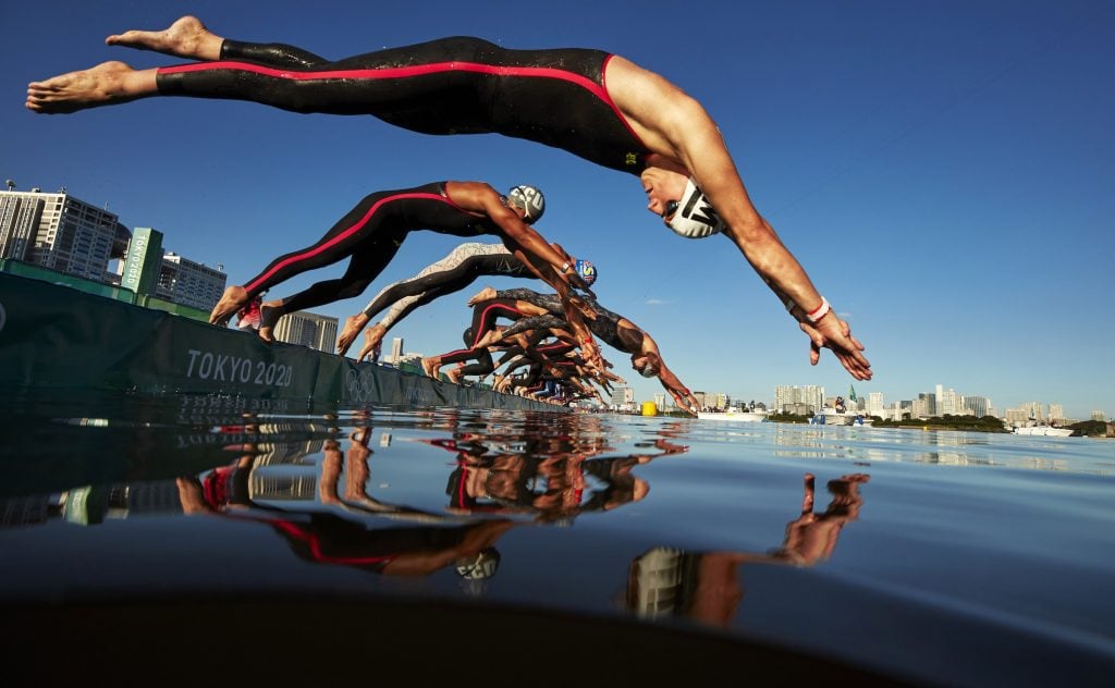 A low angle photograph of a row of swimmers diving into the water off a platform reading 