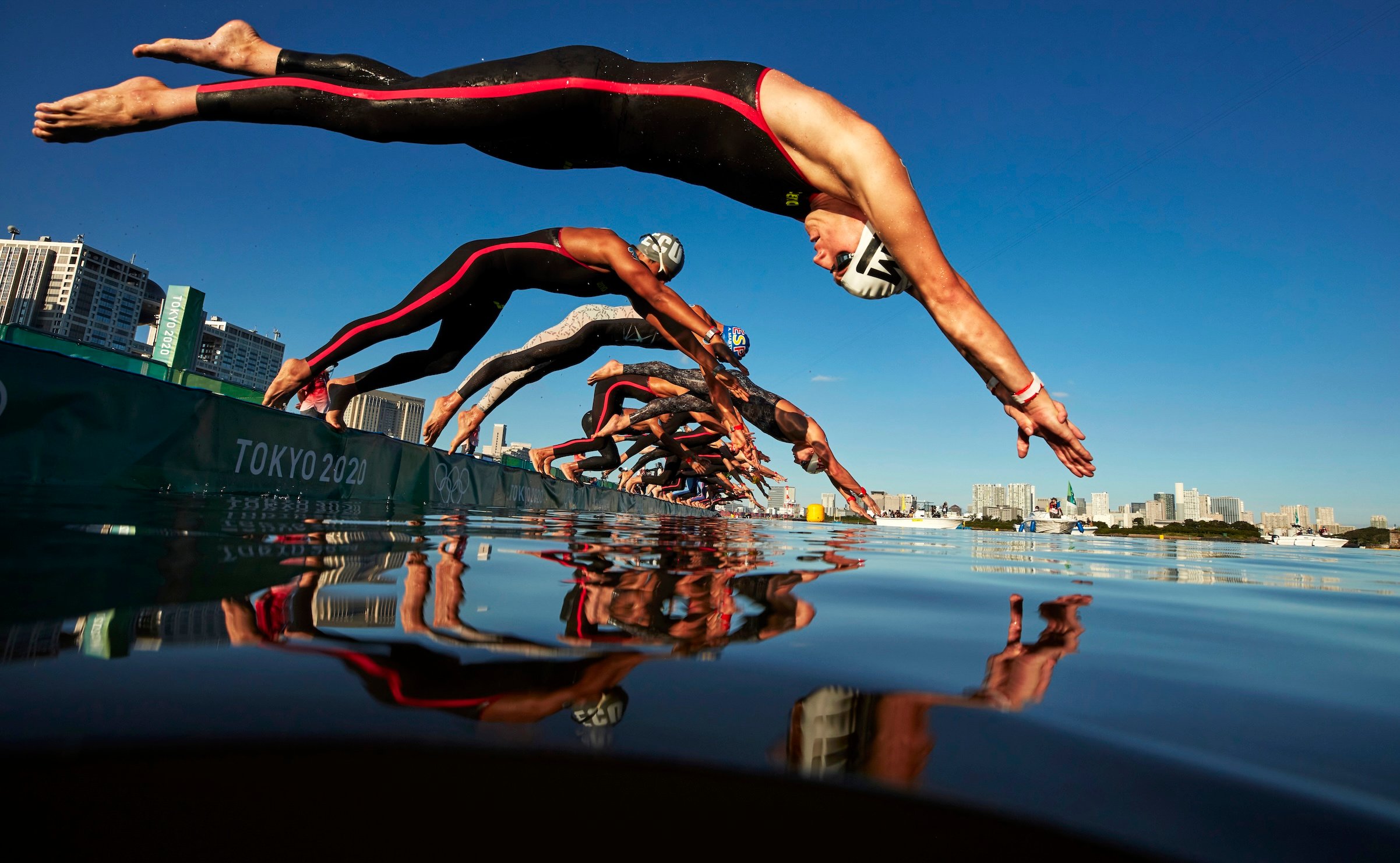 A low angle photograph of a row of swimmers diving into the water off a platform reading "Tokyo 2020." The sky behind them is a bright blue with city buildings behind them, and their reflection is mirrored in the water below.