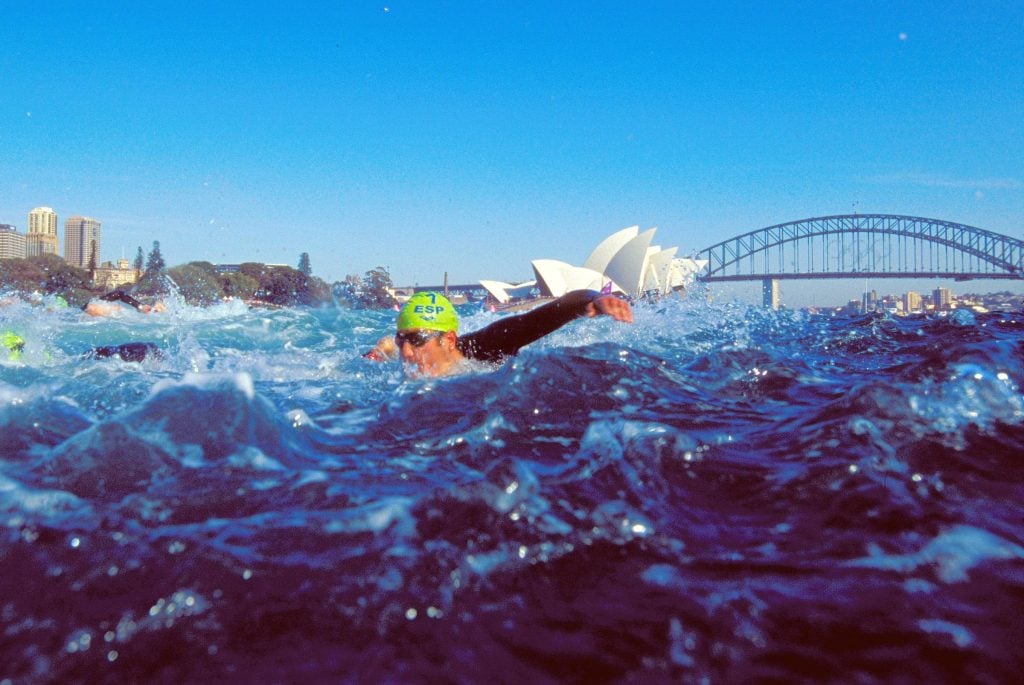 Competitors in action during the swim leg of the Men's Triathlon at the Opera House during the 2000 Sydney Olympic Games. It's a low angle photo, with an athlete in a Spain swim cap approaching the camera.