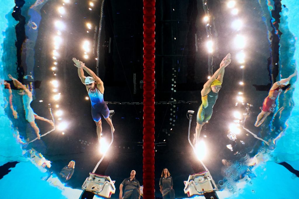 Adam Pretty used an underwater robotic camera to take this photo of U.S swimmer Katie Ledecky and her Australian rival Ariarne Titmus competing in the Women's 400m Freestyle Heats on day one of the 2024 Paris Olympic Games. The wide angle shot shows the black ceiling and lights up above, and the two swimmers, divided by a red lane line, diving into the pool. On the periphery of the image stand the coaches, and the other athletes in the outer lanes. 