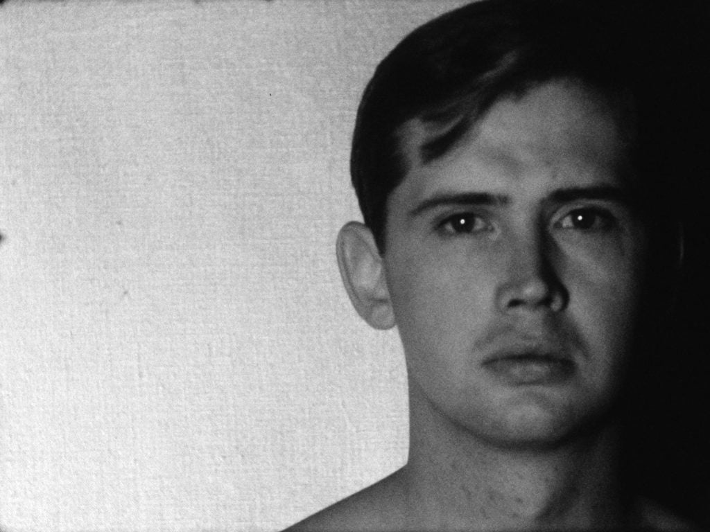 A black and white Andy Warhol film still of a handsome young man's head, facing the camera on the right of the frame, with the right side cast in shadows.