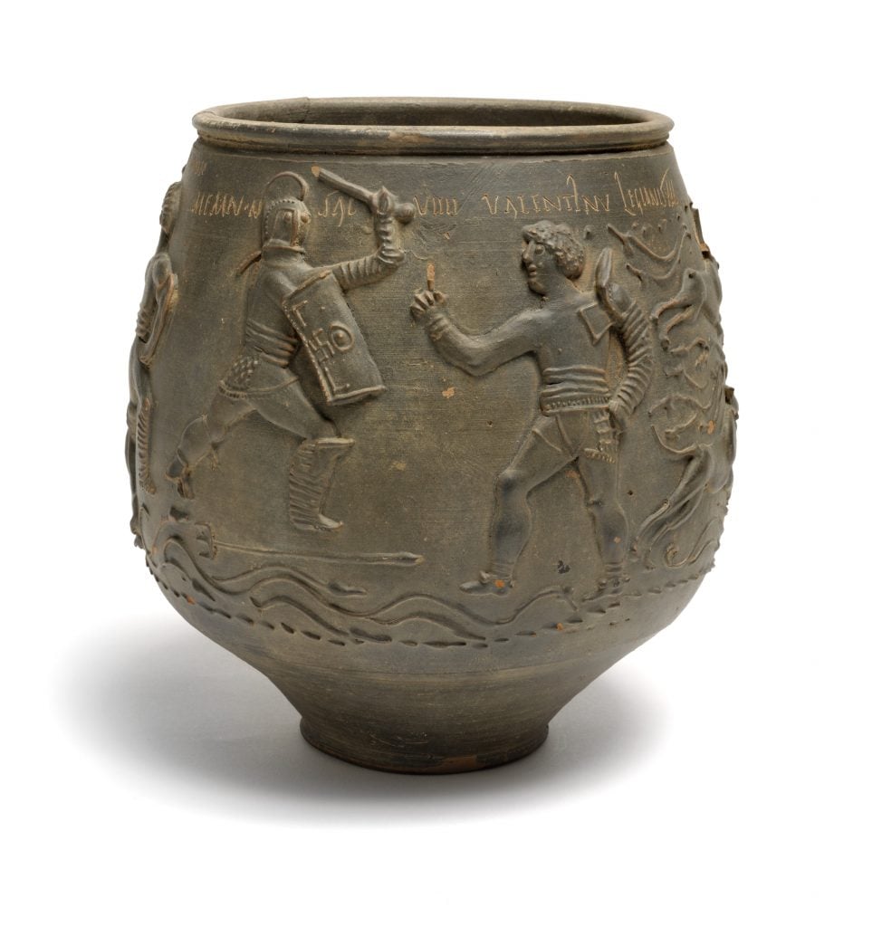 a metal vase with figures clearly carved onto it in relief, in the bit we can see there are two armoured people fighting