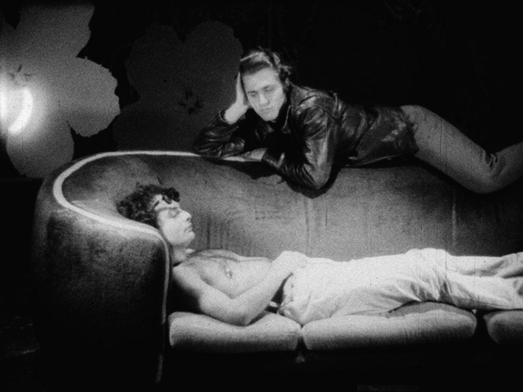 A scene from "Couch," Andy Warhol's sexually explicit black and white film featuring various visitors to his factory on his couch, often engaging in sexual activity. Here, one man in a leather jacket lies across the back of the couch looking down at a shirtless man lying below him, hands resting on his crotch. 