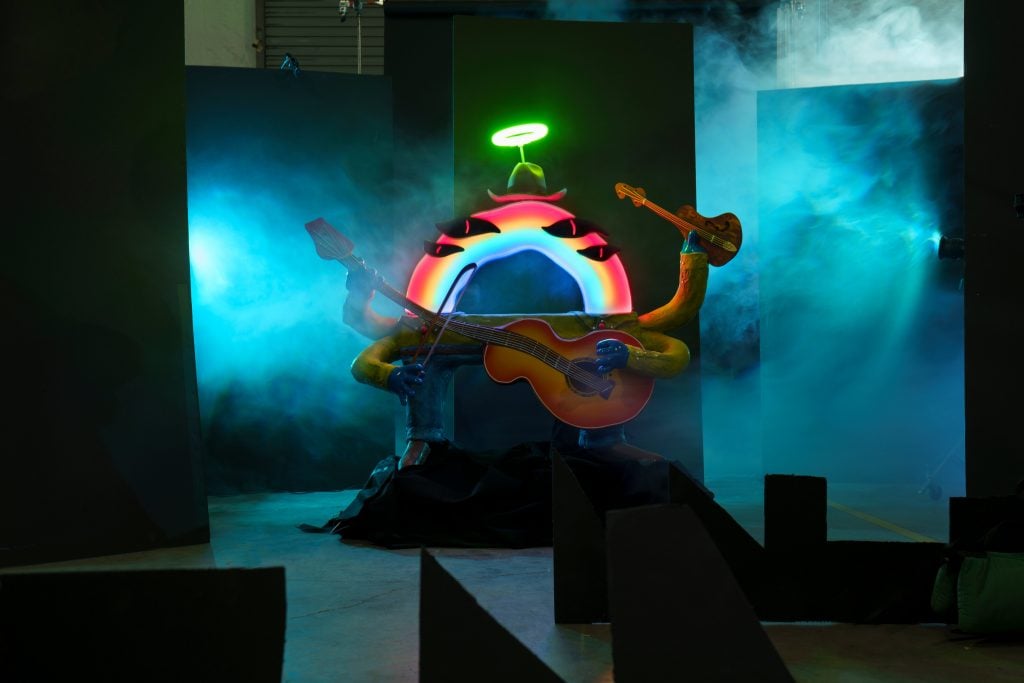 An anthropomorphic sculpture of a rainbow on a darkened stage at the Cowboix Hevvven bar at Meow Wolf Houston, with atmospheric blue fog wafting into the frame.