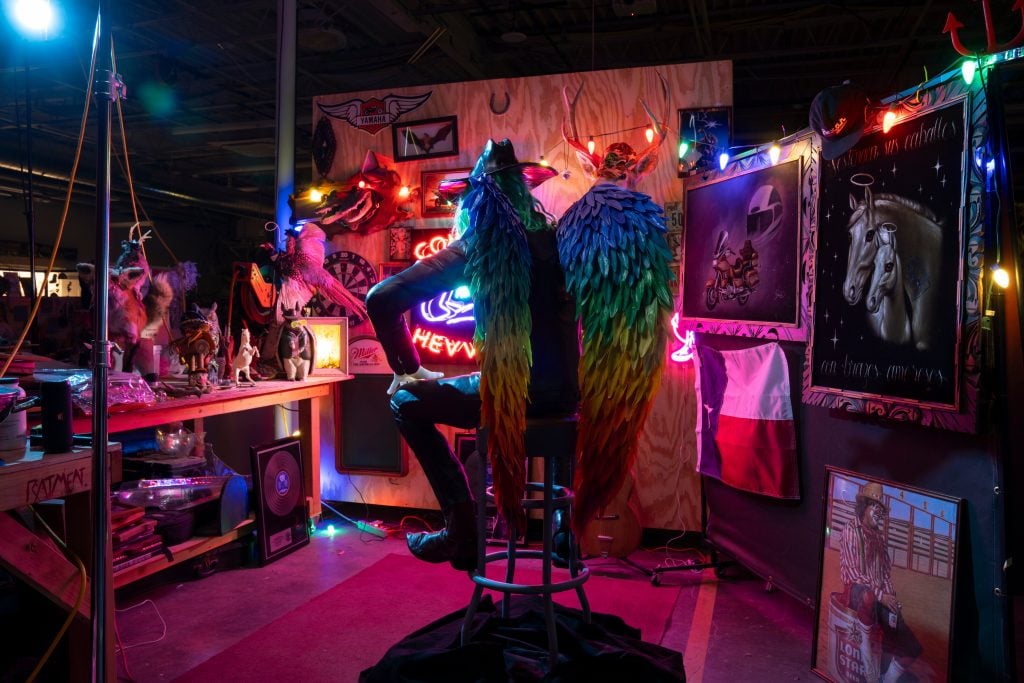 An installation view of the new Cowboix Heavvven bar at Meow Wolf Houston, a darkened interior lit by neon lights with artwork on the wall and a sculpted figure with rainbow angel wings and a cowboy hat sitting on a stool facing away from the camera.