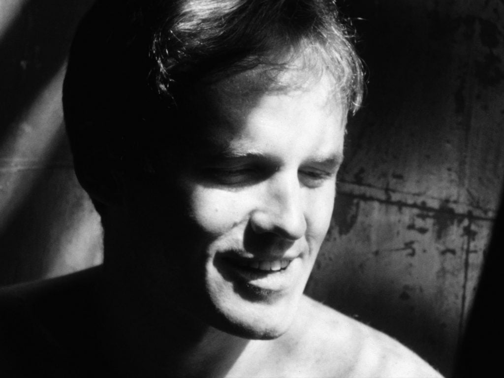 A black and white Andy Warhol film still of a young man's head, looking to the right with shadows across the left side of his smiling face.