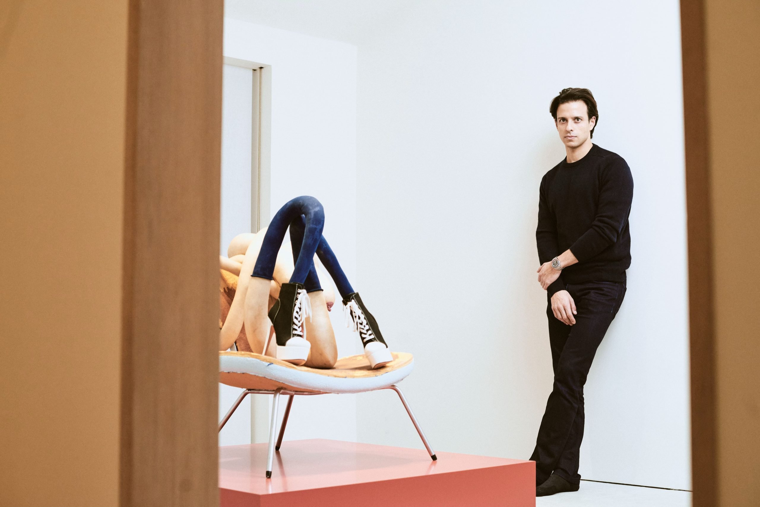 A man in dark hair and dark outfit posing in an art gallery.