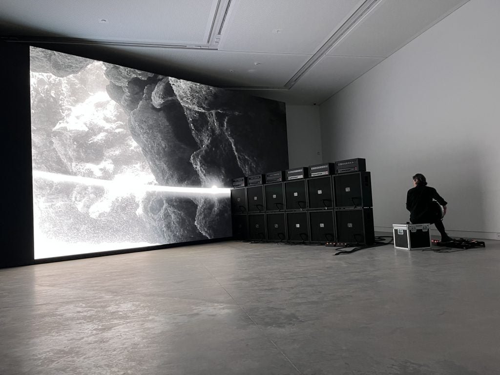 A big screen in black and white in a room, with loudspeakers installed and a man playing the guitar