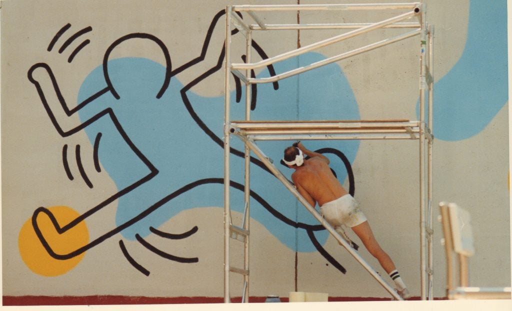 a shirtless man leans against scaffolding to paint a giant outline as part of a mural