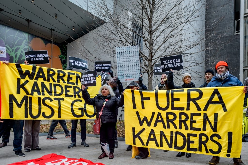 protestors stand with large yellow and black banners outside of a museum in new york