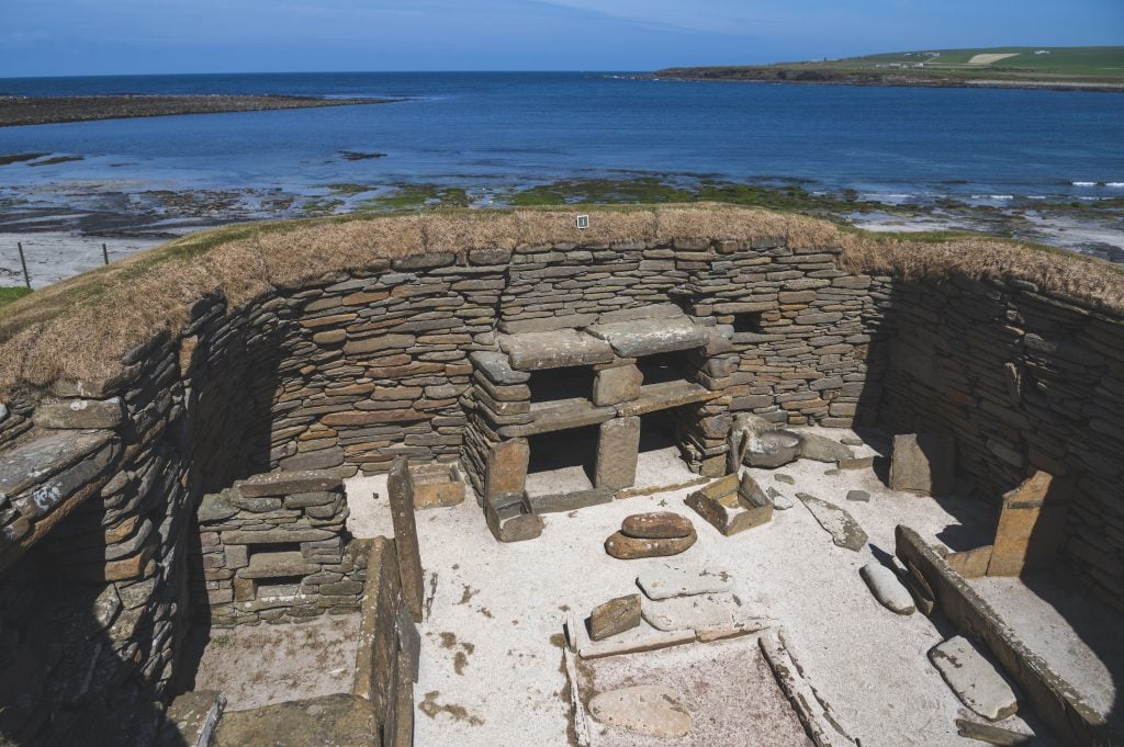 A view of one of the houses at Skara Brae, Orkney, the sea is in the background and we can see the tight layering of stones as well as internal features of the house such as hearth.