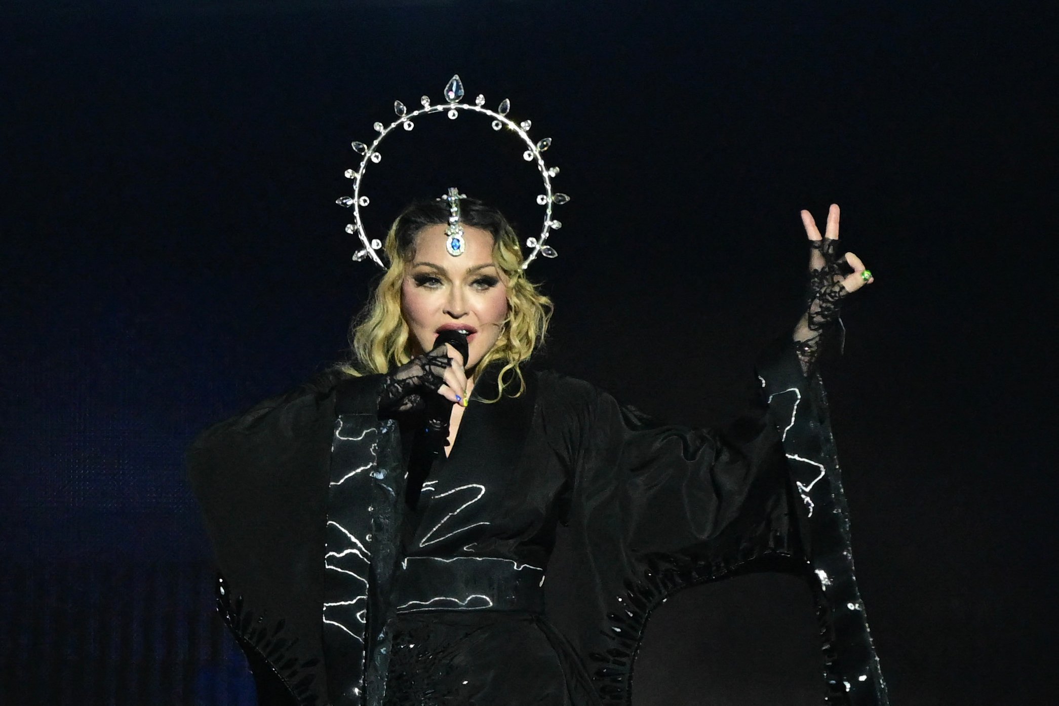 Pop star Madonna stands onstage with a silver crown over head with a black background behind her.