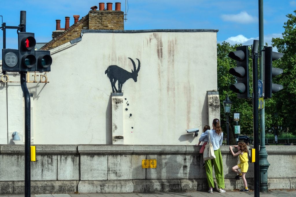 The side of a London house is painted white and has a small ledge on it with an image of a mountain goat stencilled onto it, giving the impression that it is balancing on the ledge, looking down, with boulders falling off where its hooves are.