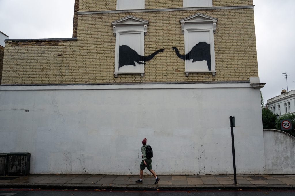 the side of a classic London house has two windows in a brick facade with no actual glass window in them, and inside each is a stencilled image of an elephant, they are turning towards each other and reaching out with their trunks