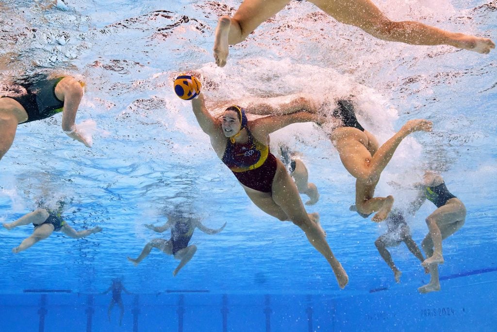 Adam Pretty used an underwater robotic camera to photograph Paula Leiton of Team Spain with the ball in the Women's Gold Medal match between Team Australia and Team Spain on day fifteen of the 2024 Paris Olympic Games. She is seen from below, the blue and yellow ball in her right hand, her face underwater grimacing. The other players around her all have their heads above water, with only their legs, arms and torsos visible in the shot.