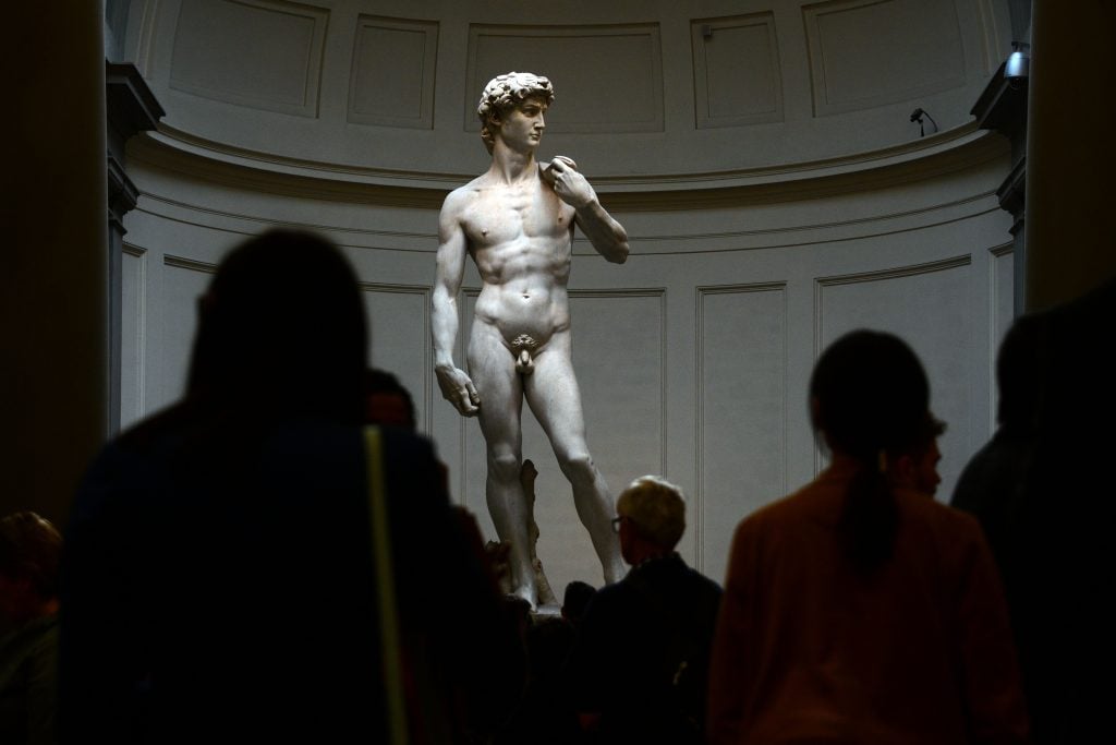 the backs of a group of tourists are seen in shadow as they look at the white marble statue of david