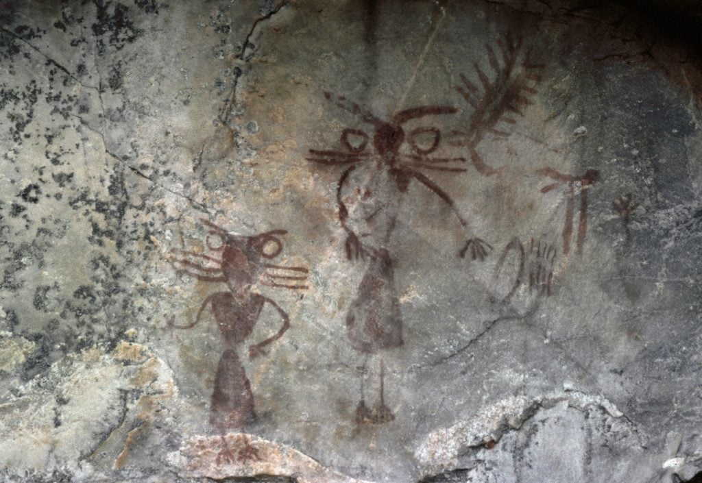 a photo of ancient rock paintings. two figures in dark red paint are seen on a grey cave wall