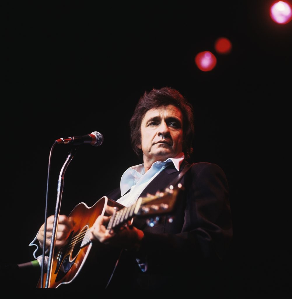 singer Jonny Cash onstage playing guitar, he is stood in front of a microphone with red lights behind him