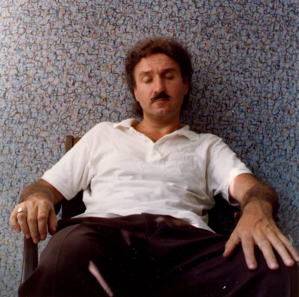 A photograph of Henry Orlik in white shirt and black pants seated in a chair with his eyes closed before a colorfull wall painted with a great number of tiny, tight spirals