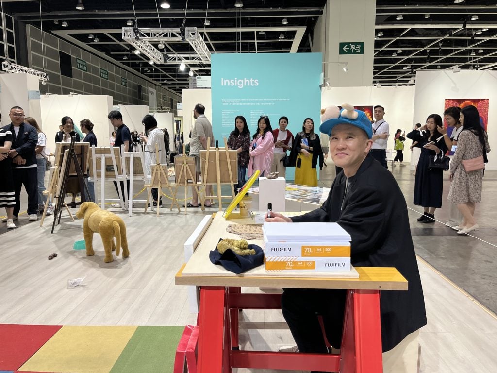 An East Asian man in a blue cap sitting a colourful desk, with a long queue at the back in an exhibition hall 