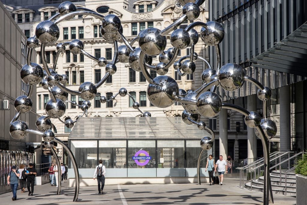 a large metal reflective structure that looks like beads threaded on a string that rises up and floats in twisted turns in the air above a tube station entrance in a very urban setting