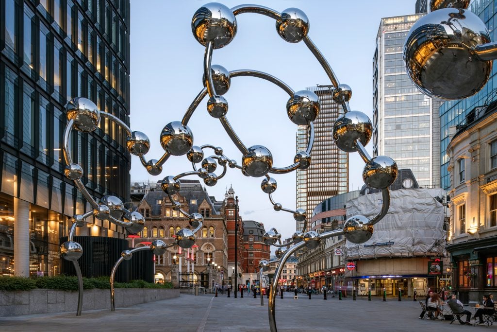 a large metal reflective structure that looks like beads threaded on a string that rises up and floats in twisted turns in the air in a very urban setting