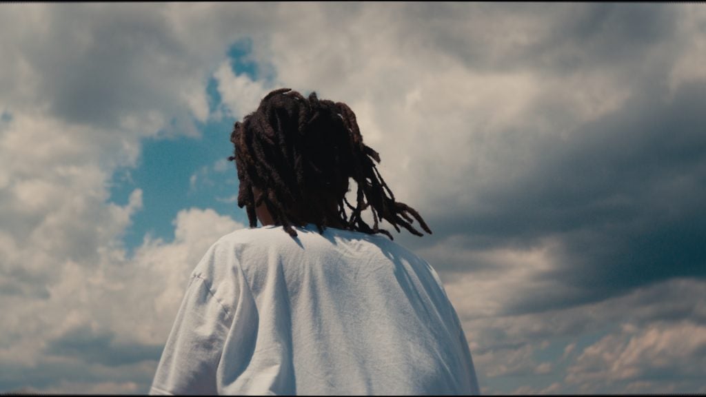 A Black woman with dreadlocks in a white robe, seen from behind from the torso up, looks up at clouds in a blue sky.