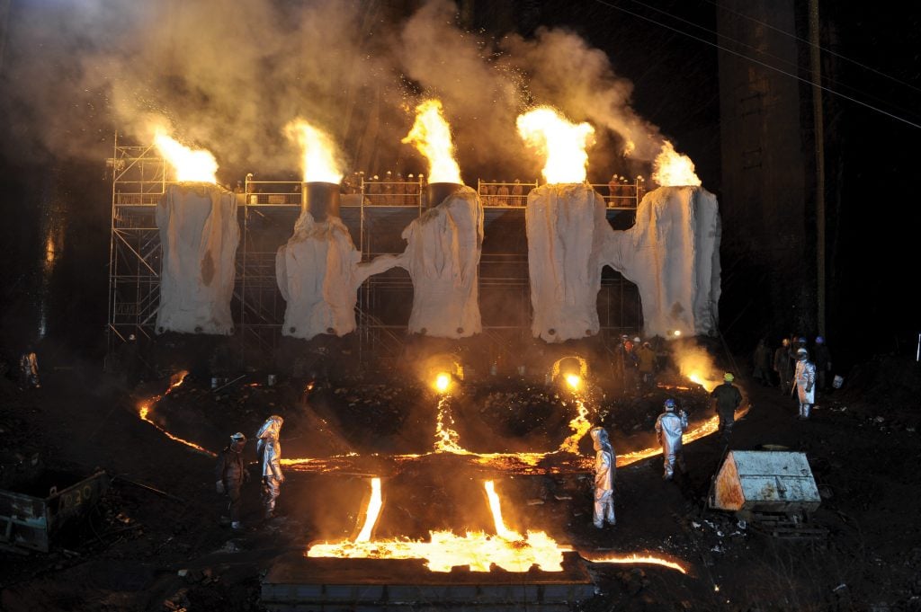 men in fire proof suits walk around a factory with pipes belching flames and liquid molten steel flowing 