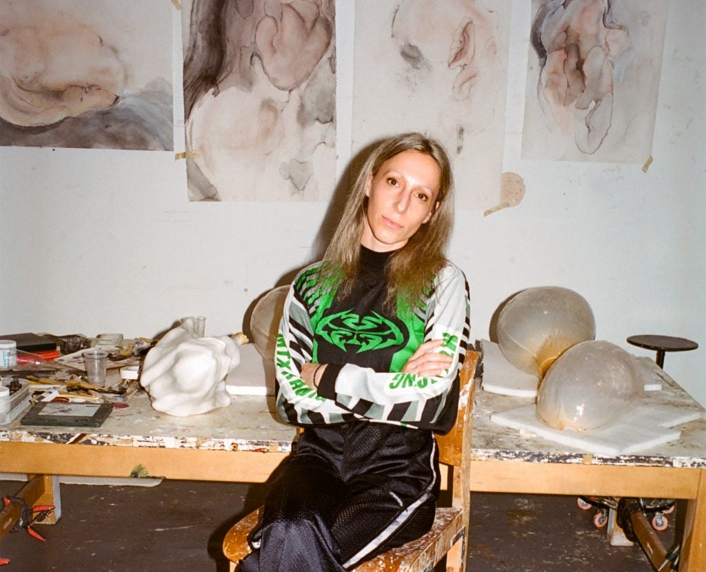 The artist Ivana Bašić sit with her arms crossed in her studio, leaning against a studio table.