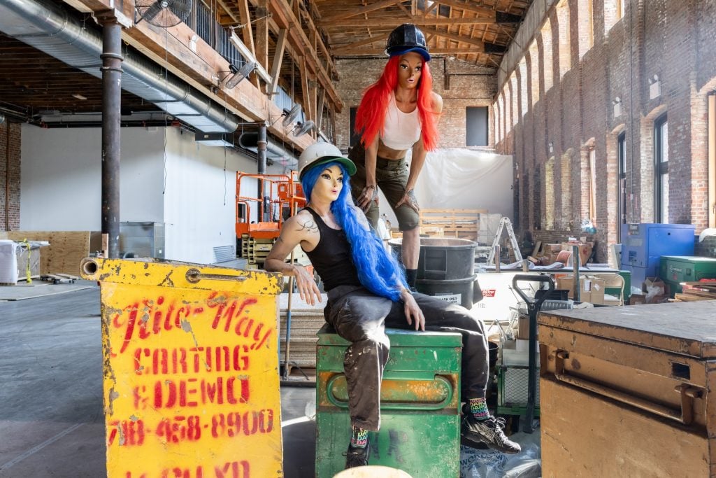 Two women in tank tops and construction hats, wearing mannequin masks, pose amid construction materials in the sunlight great hall at Brooklyn's Pioneer Works. The mask is the signature of the performance artist Narcissister, who will stage a new performance at the Red Hook cultural center.