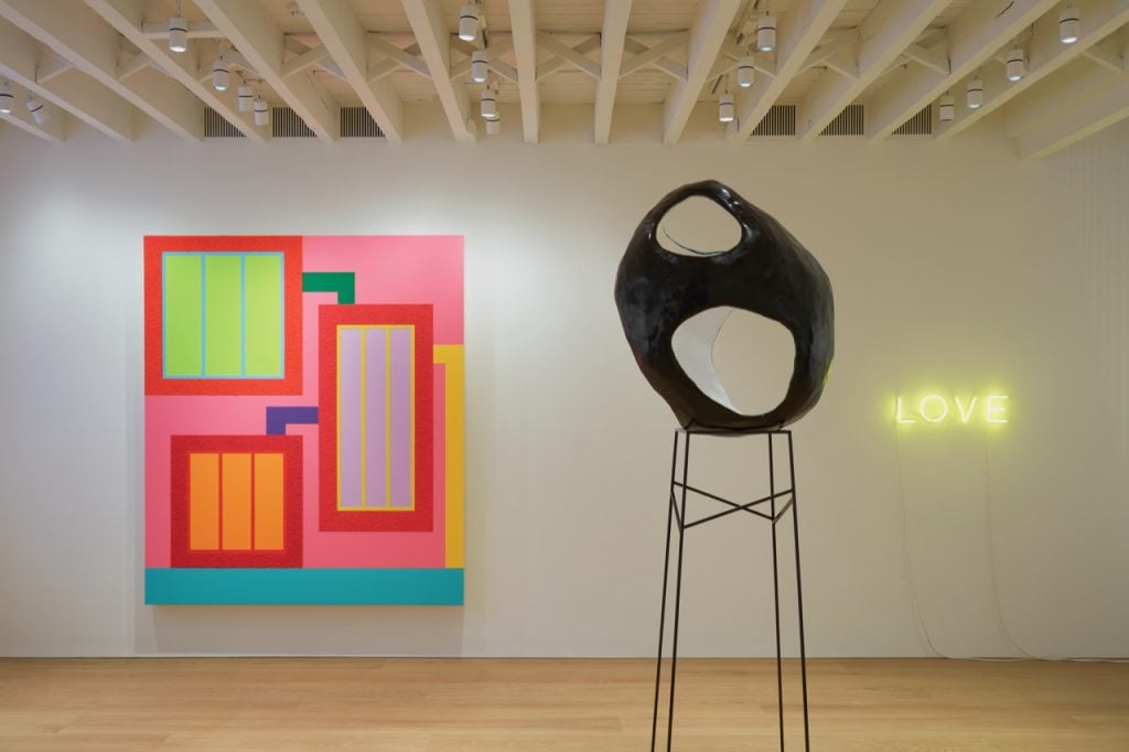 A colourful painting, a sculpture sitting on a steel frame, and a yellow neon installation on the wall. 