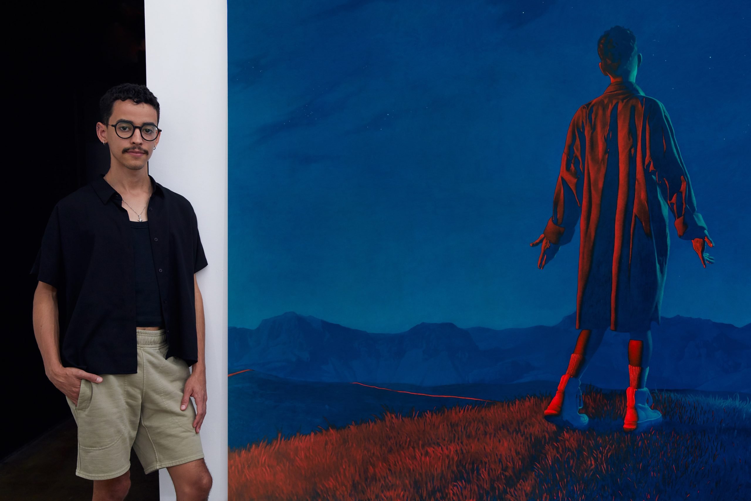 the artist Esaí Alfredo stands in front of his painting depicting a male form standing on a scenic butte