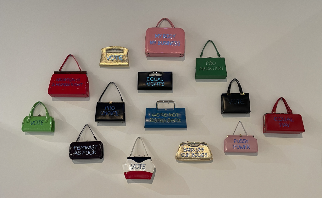 Works from Michele Pred's series "The power of the wallet" to see in "In action 2024." A group of colorful vintage handbags hangs on a white wall. The bags carry feminist messages such as "Pussy Power" And "Prohibitions from our body" written on it with electroluminescent wire.