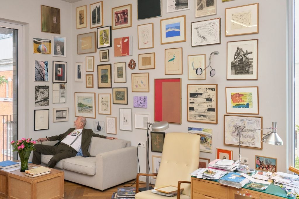 This image depicts a man sitting on a grey couch with lots of artworks on the wall