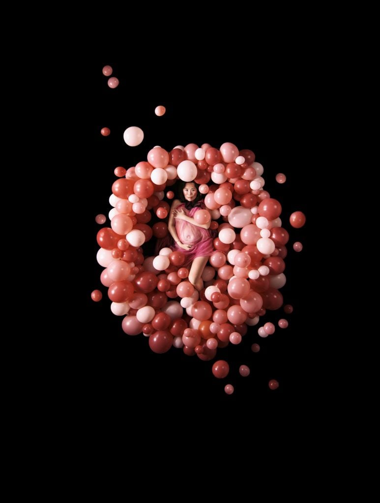 This photo depicts a woman surrounded by many pink balloons against a dark background