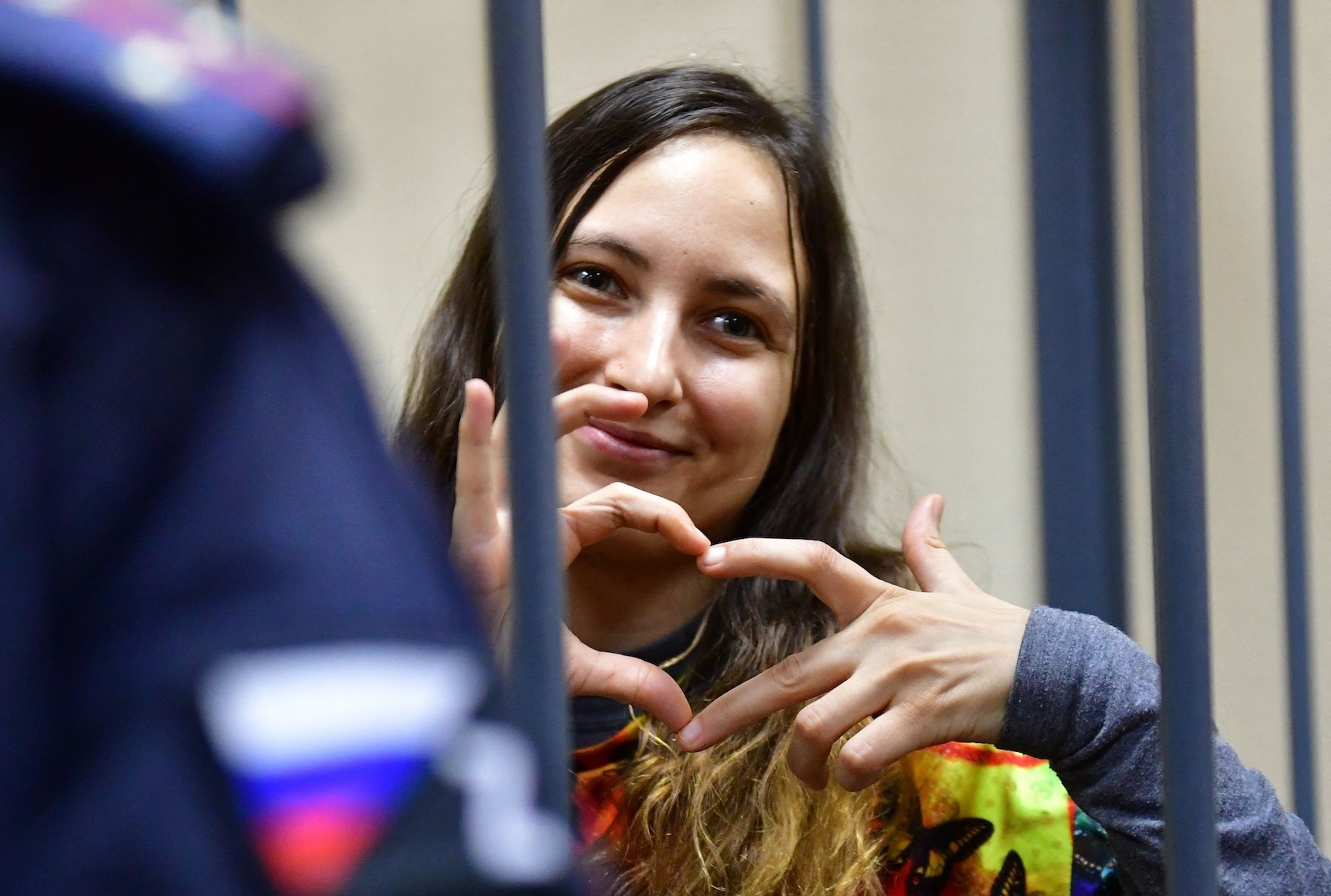 A woman, artist Sasha Sasha Skochilenko, behind bars makes a heart shape with her fingers