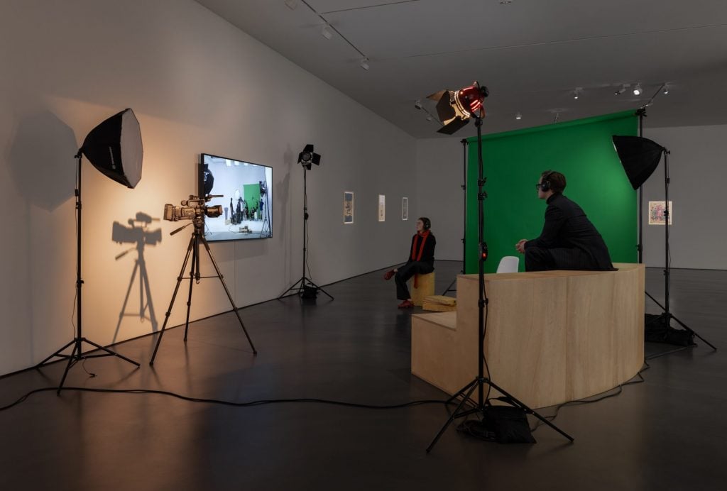 This image shows audience members sitting in front of a TV screen watching a video at a gallery space.