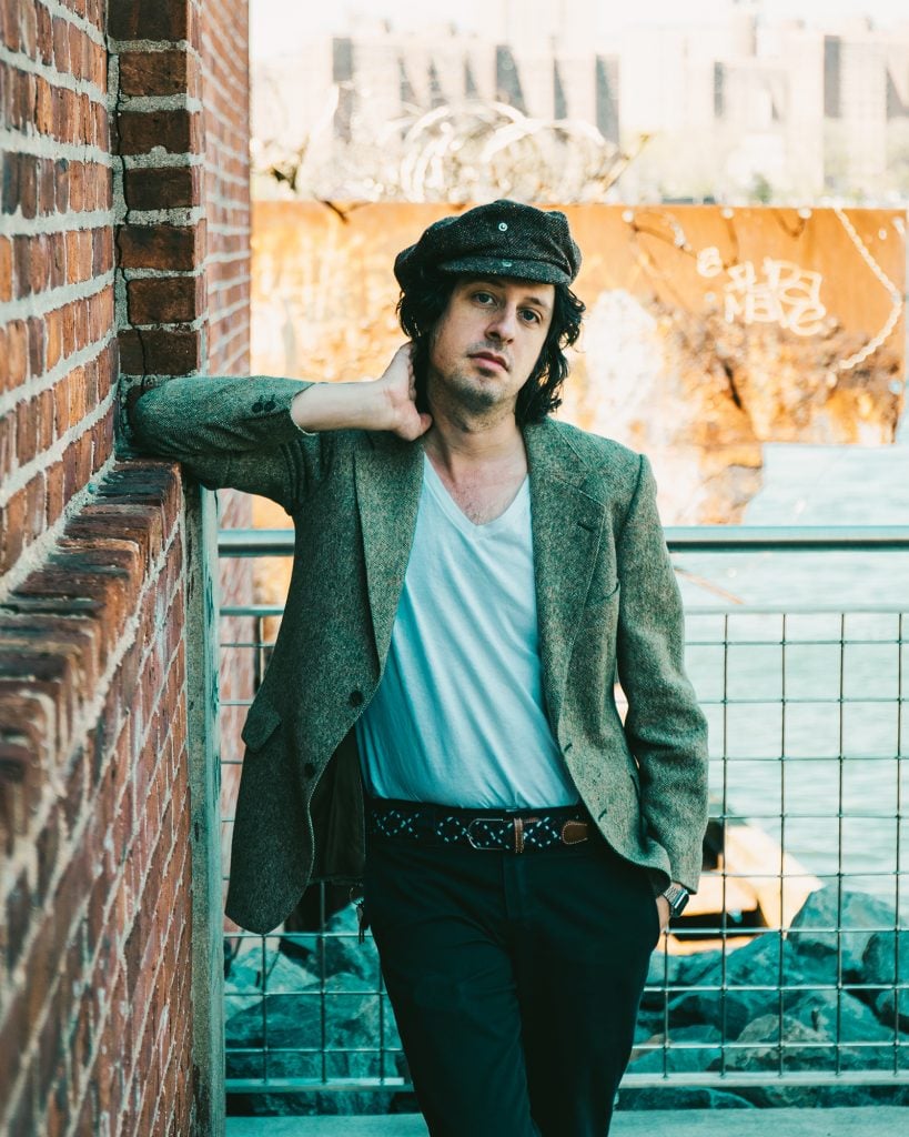 A man, musician Adam Green, posing on a balcony in a cap, loose-t-shirt and blazer