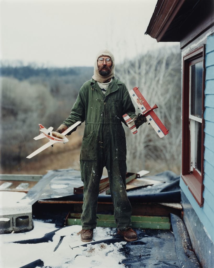 A bearded man in a rural winter landscape holds two model planes