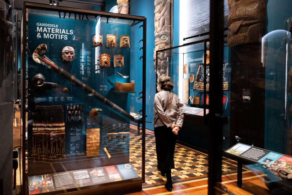 an interior gallery space with blue walls and glass cases containing Native American artifacts, a woman can be seen walking through