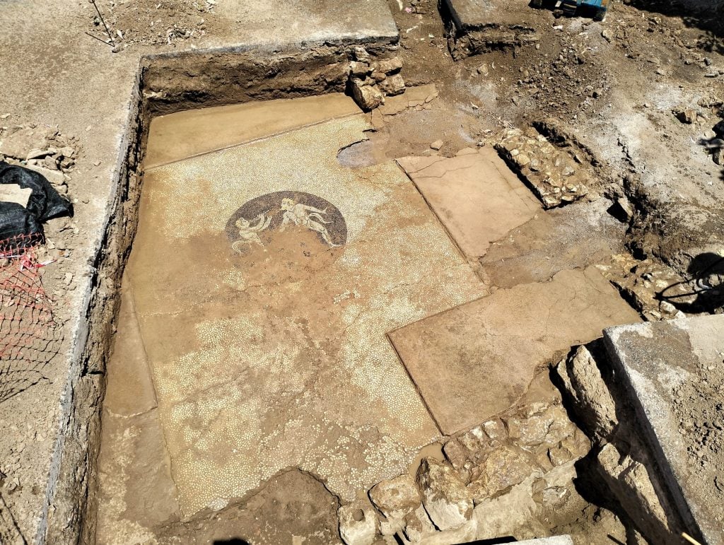 An ancient Greek mosaic depicting two dancing satyrs seen under a layer of dirt on a crumbling floor