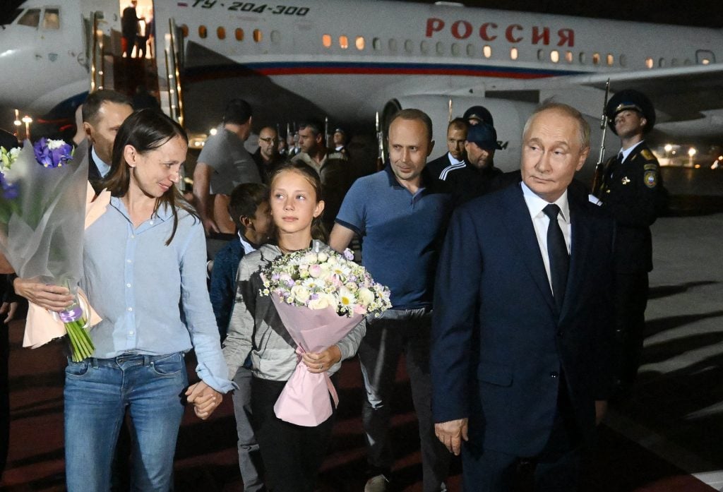 A woman, Anna Dultseva, clutching the hand of a young girl beside Russian prime minister Vladimir Putin. An airplane sits behind them