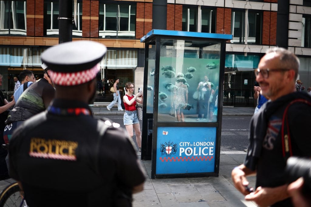 a street scene in London in daylight, we can see the back of a uniformed police officer and a Metropolitan police box with glass windows. this glass surface with a school of piranha fish in shades of semi-transparent, shimmery blue pasted onto it