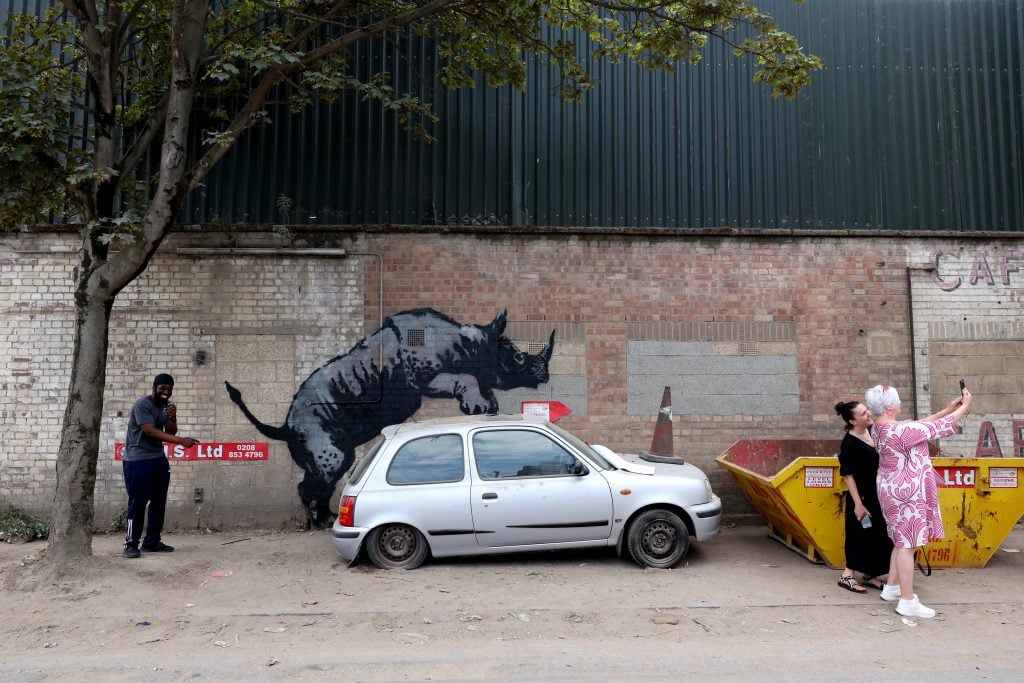 a graffiti image of a grey rhinosceros appears to be mounting from behind a beaten up small grey car with a horn on its bonnet outside on the side of a quiet road. There is a skip next to the car, two people are taking a selfie with the artwork and another is pointing at it and laughing