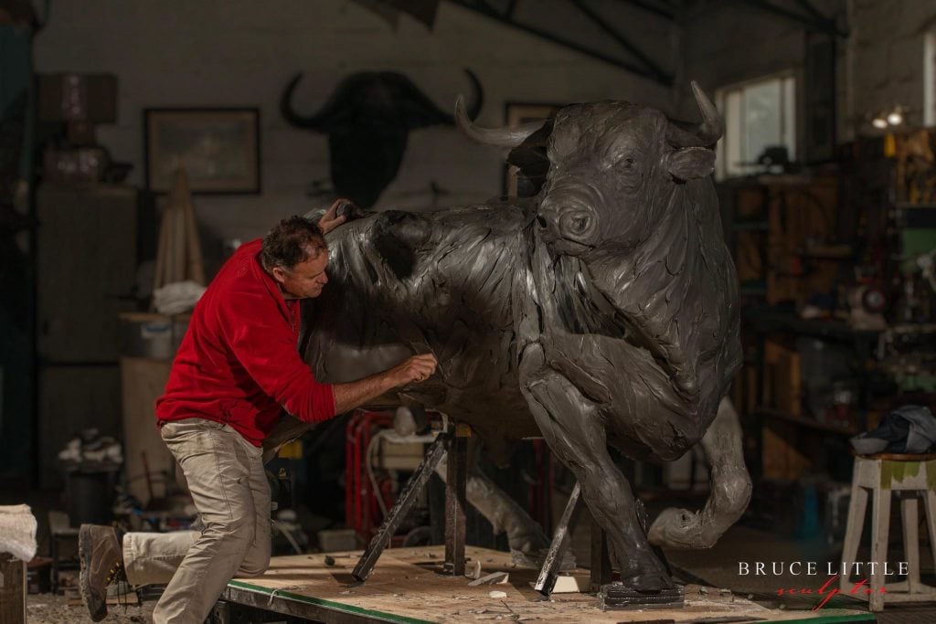 Artist Bruce Little in his studio wearing a red long sleeve shirt and khakis working on a life-size sculpture of a bull in bronze.