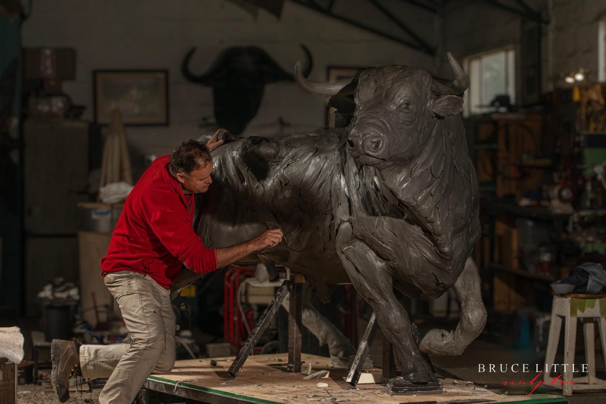 Artist Bruce Little in his studio wearing a red long sleeve shirt and khakis working on a life-size sculpture of a bull in bronze.