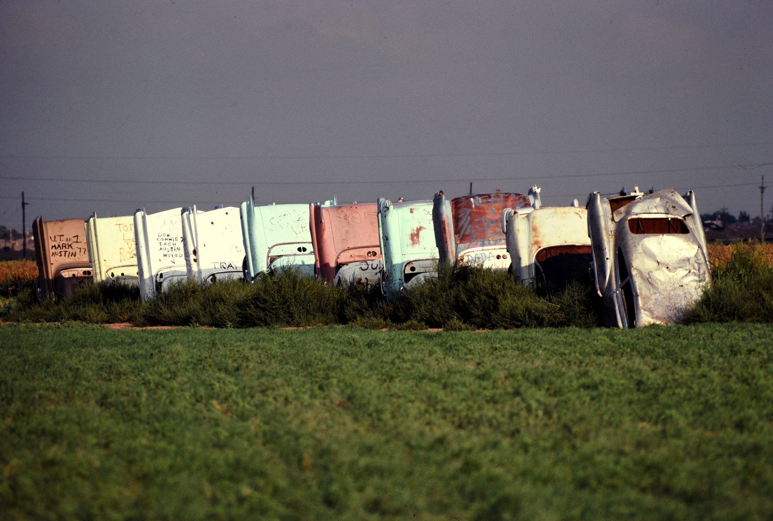 A photograph of ten Cadillac cars sticking out of the ground as part of Cadillac Ranch, with green grass in the foreground and dark grey skies in the background