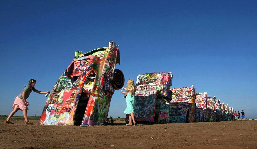 A photo of two girls spray painting a row of Cadillac cars half buried in desert dirt as part of the Cadillac Ranch under a blue sky